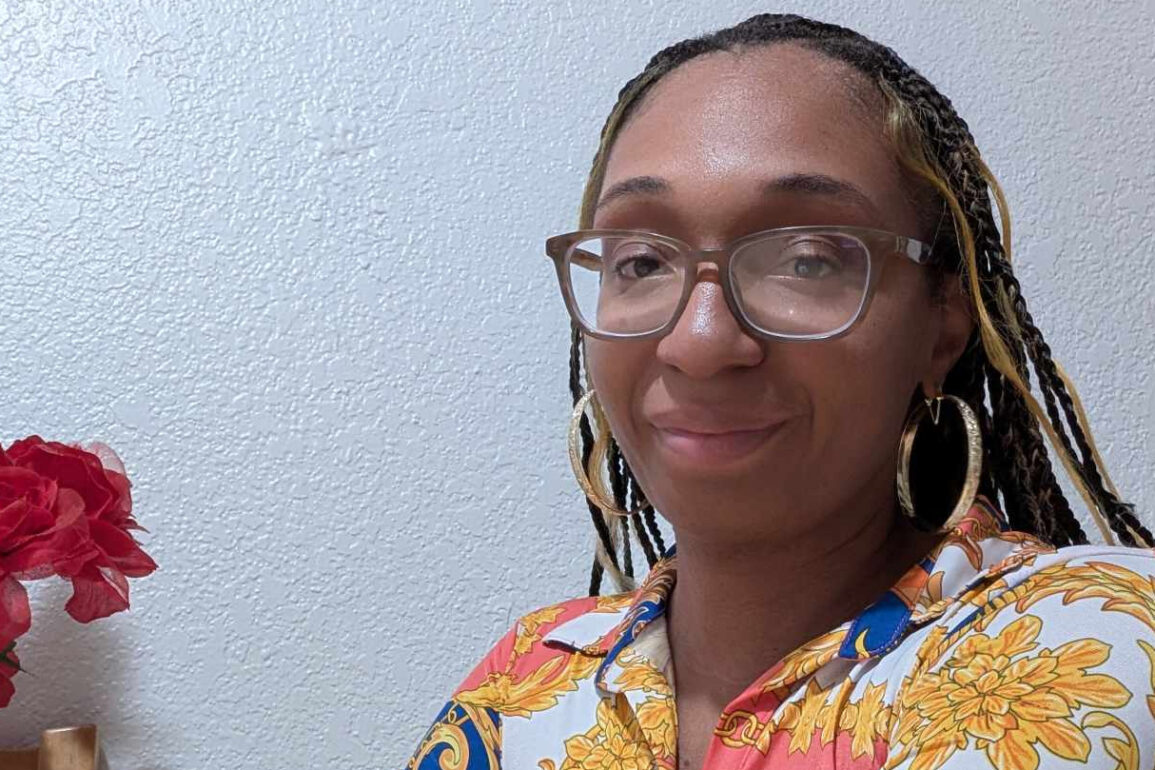 Photo of Jessica 'Nooni' Noble, Editor-in-Chief of Sports and More Sports, wearing glasses and a vibrant patterned shirt, smiling against a textured white wall with red and pink flowers in the foreground. Photo featured in the Sports And More Sports article, "Jessica “Nooni” Noble: From Gamer to Game Changer in Sports Journalism."