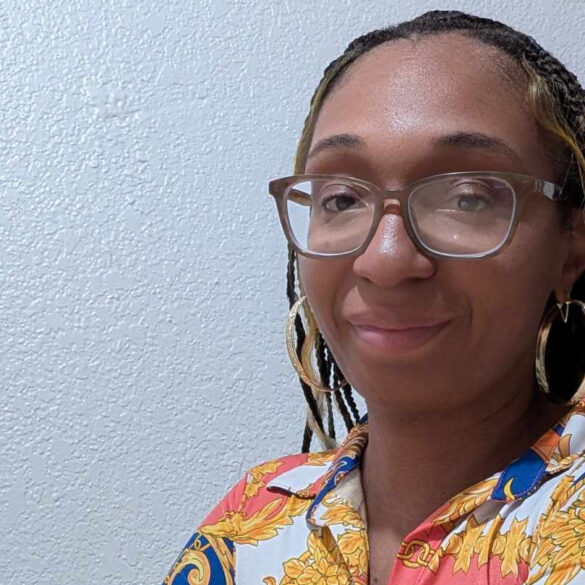 Photo of Jessica 'Nooni' Noble, Editor-in-Chief of Sports and More Sports, wearing glasses and a vibrant patterned shirt, smiling against a textured white wall with red and pink flowers in the foreground. Photo featured in the Sports And More Sports article, "Jessica “Nooni” Noble: From Gamer to Game Changer in Sports Journalism."